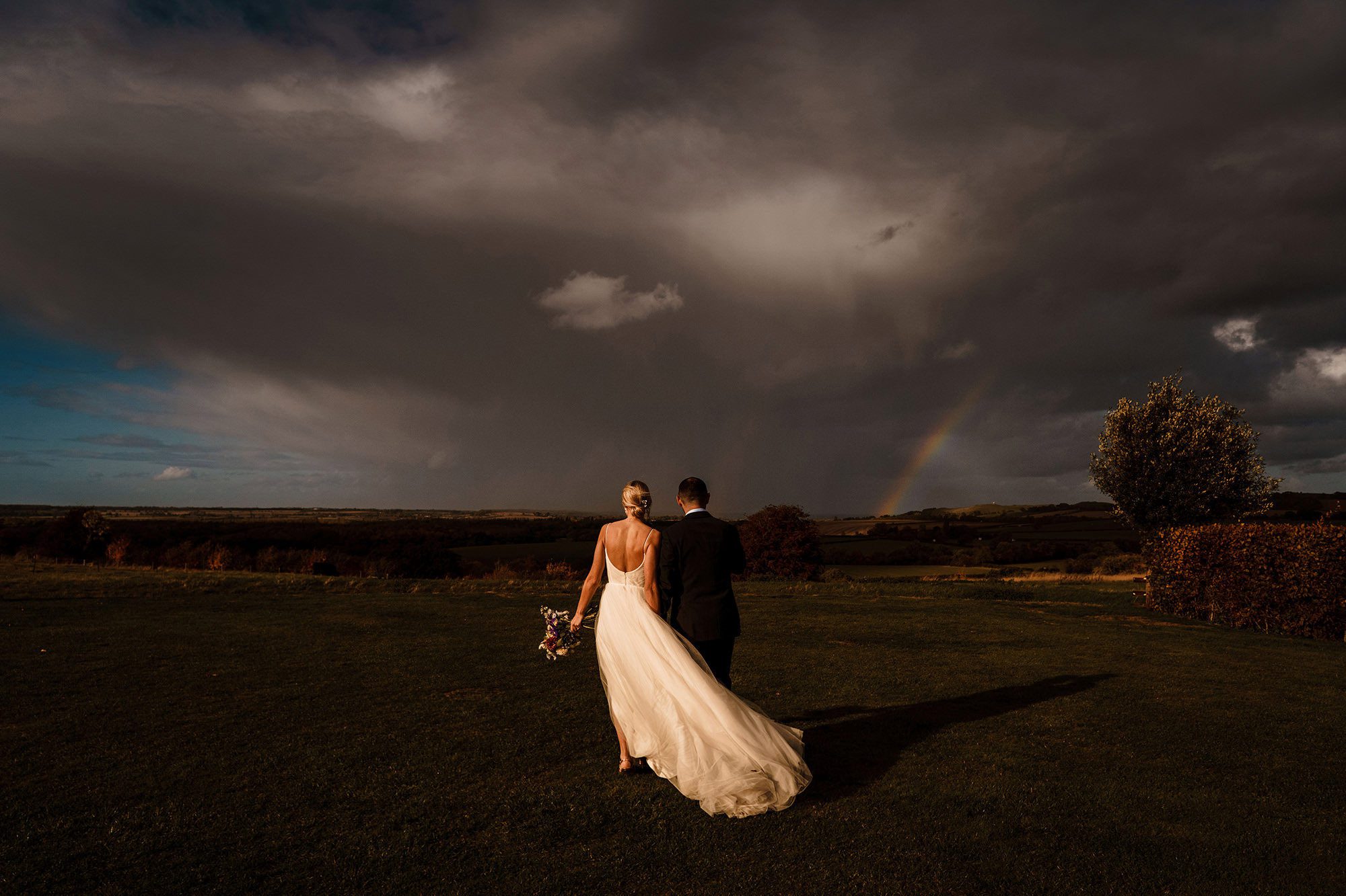 Bride and groom pose as a rainbow pierces distant storm.